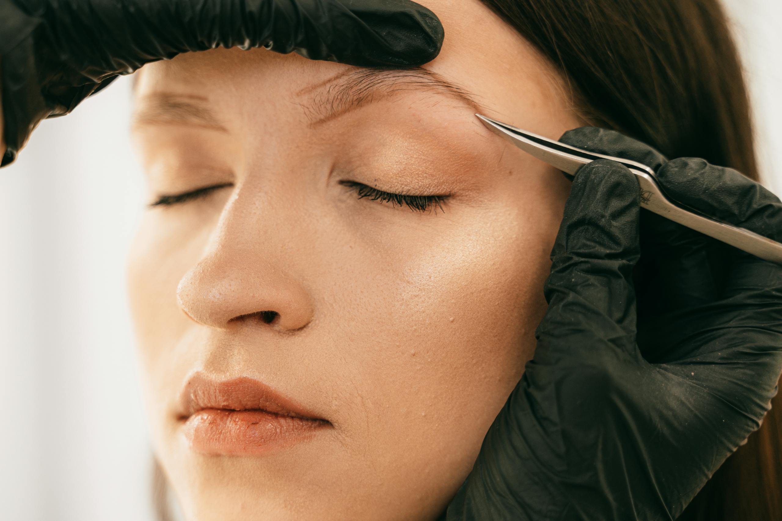 Detailed image of woman getting eyebrows plucked with tweezers by a beautician wearing gloves.